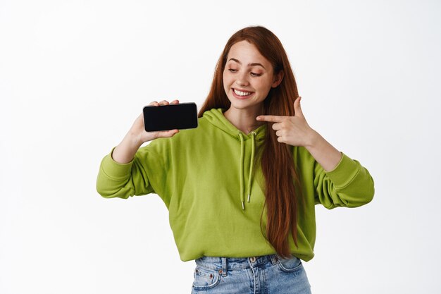 Chica pelirroja alegre apuntando a la aplicación de teléfono móvil horizontal, mostrando la pantalla de la tienda en línea y sonriendo o videojuego en blanco