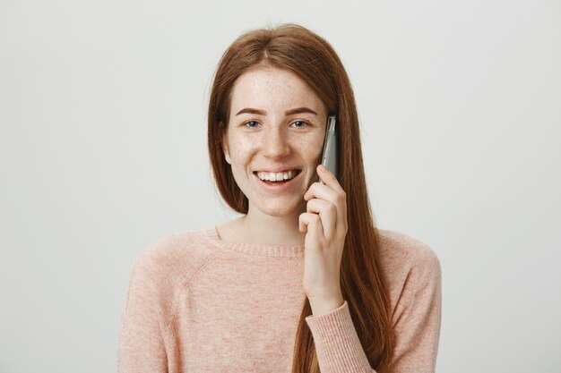 Chica pelirroja alegre con pecas, sonriendo y hablando por teléfono sin preocupaciones