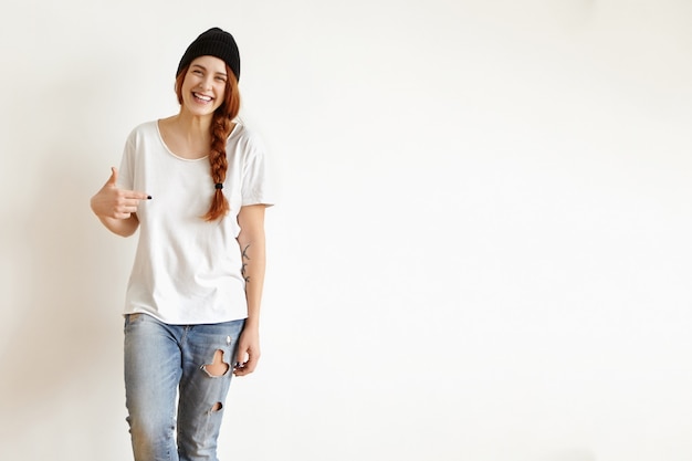 Foto gratuita chica pelirroja alegre con peinado trenzado con sombrero negro y jeans raídos, señalando con el dedo su camiseta blanca de gran tamaño