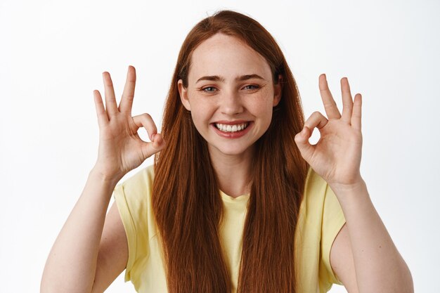 Chica pelirroja alegre con piel limpia y dientes blancos, sonriendo y mostrando el signo de OK OK, alaba la buena elección, de pie en blanco