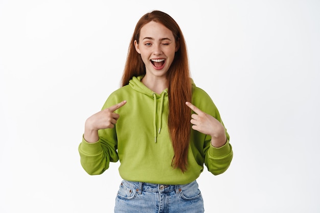 Chica pelirroja alegre, señalando a sí misma y diciendo que soy, guiñando un ojo y sonriendo feliz, de pie con ropa casual en blanco.