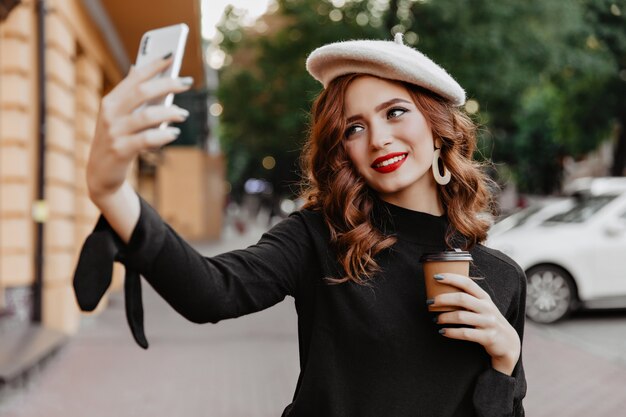 Chica pelirroja alegre sosteniendo una taza de café y posando al aire libre. Joven francesa despreocupada haciendo selfie en la calle.