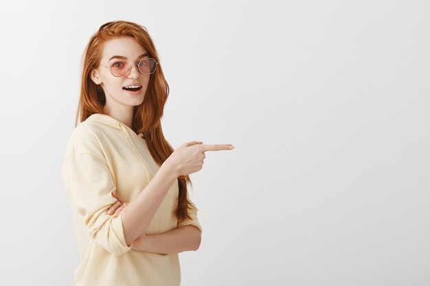 Chica pelirroja atractiva con estilo en gafas de sol apuntando con el dedo a la derecha en copyspace