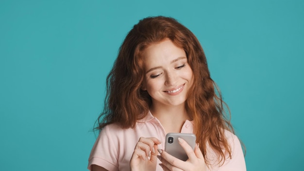 Chica pelirroja bastante encantadora usando felizmente el teléfono inteligente sobre fondo colorido