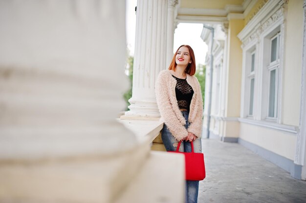 Chica pelirroja con bolso rojo posado cerca de casa vintage