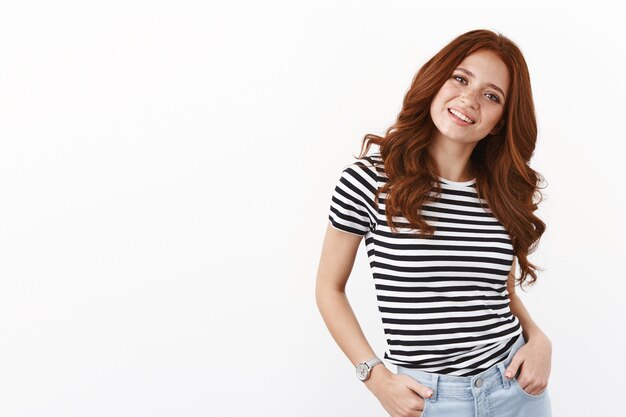 Chica pelirroja caucásica moderna alegre con pecas en camiseta a rayas, tomados de la mano en los bolsillos, inclina la cabeza con alegría, curiosa, sonriente, mirada entusiasta de la cámara, hablando casualmente, pared blanca