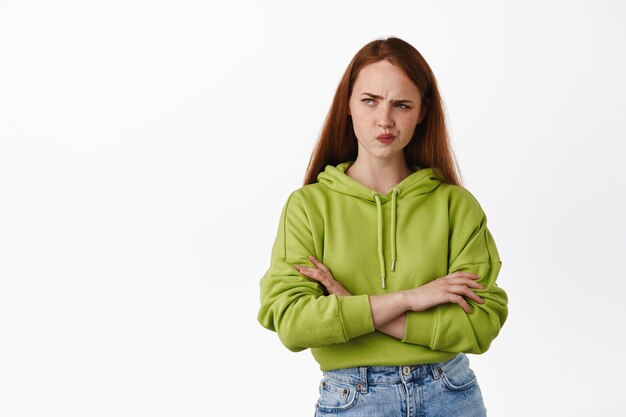 La chica pelirroja complicada parece perpleja, frunce el ceño y se enfurruña mientras piensa, mira a un lado y toma una decisión, siente incredulidad sospechosa, no está segura, fondo blanco.