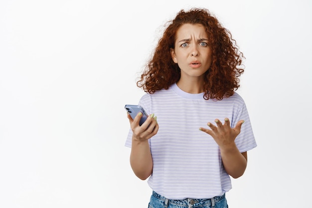 Foto gratuita chica pelirroja confundida que parece desconcertada, sostiene un teléfono inteligente, no puede entender algo en el teléfono, de pie sobre fondo blanco