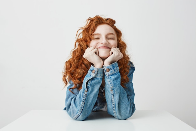 Foto gratuita chica pelirroja contenta sonriendo regocijo con las manos en las mejillas sentado en la mesa.