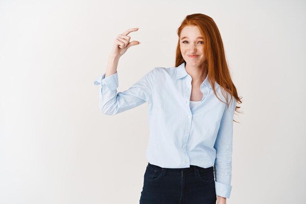 Chica pelirroja decepcionada con camisa azul mostrando algo pequeño y haciendo una mueca incómoda juzgando algo pequeño o pequeño de pie sobre fondo blanco