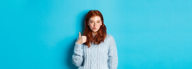 Foto gratuita chica pelirroja descarada en suéter luciendo complacida y mostrando el pulgar hacia arriba como y de acuerdo de pie sobre azul