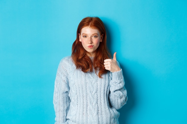 Chica pelirroja descarada en suéter, luciendo complacida y mostrando el pulgar hacia arriba, como y de acuerdo, de pie sobre un fondo azul.