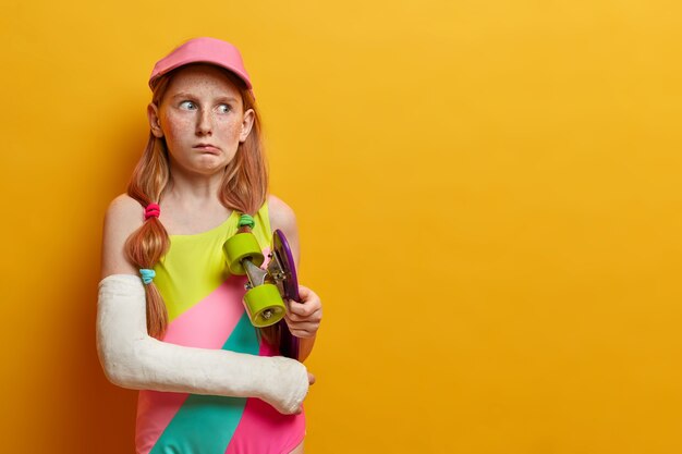 Chica pelirroja desconcertada sostiene el longboard debajo del brazo, posa con el patín y la mano rota en el yeso, tiene un día desafortunado y parece disgustada a un lado, aislada en la pared amarilla, se lesionó después de hacer un truco