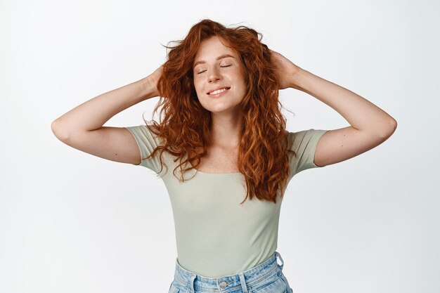 Chica pelirroja despreocupada con cabello rojo saludable y natural tomándose de la mano detrás de la cabeza y descansando acostada con una sonrisa complacida y los ojos cerrados de pie sobre fondo blanco