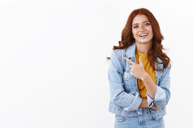 Chica pelirroja elegante con chaqueta vaquera, apuntando con el dedo hacia la izquierda y sonriendo confiada, dando consejos, sugiriendo promo, indicando espacio de copia lateral, de pie en la pared blanca alegre y decidida