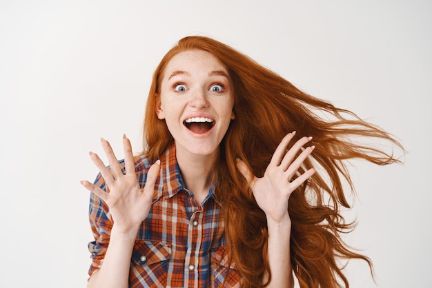 Chica pelirroja emocionada haciendo un anuncio gritando de alegría y felicidad mirando a la cámara cabello de jengibre asombrado volando en el aire fondo blanco