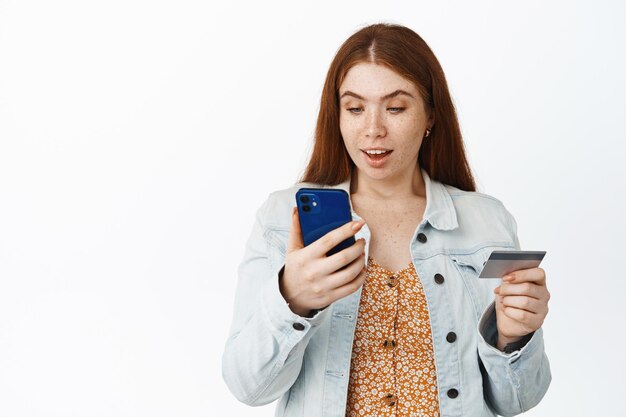 Una chica pelirroja entusiasta hace un pago en línea mirando su teléfono mientras mantiene un pedido de tarjeta de crédito en el fondo blanco de la aplicación