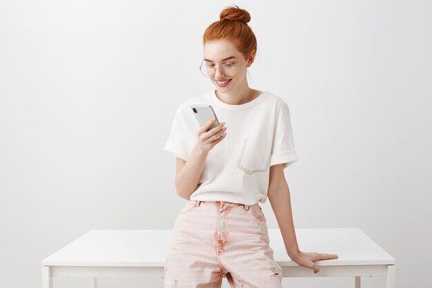 Chica pelirroja con estilo teniendo descanso, mirando el teléfono móvil complacido