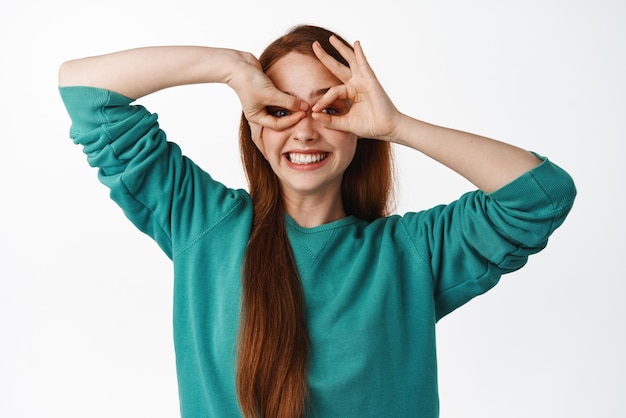 Chica pelirroja feliz mira a través de los dedos gafas binoculares sonriendo feliz y positiva haciendo máscara de superhéroe y jugando de pie sobre fondo blanco.