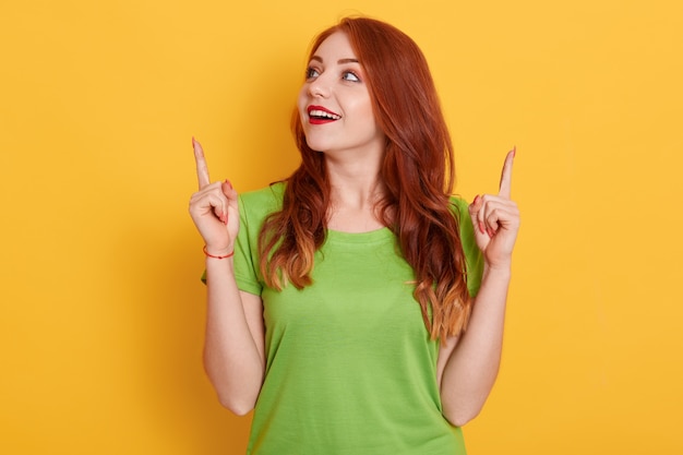 Chica pelirroja feliz sorprendida en camiseta verde casual mirando asombrado y apuntando hacia arriba con los dedos índices, posando aislado