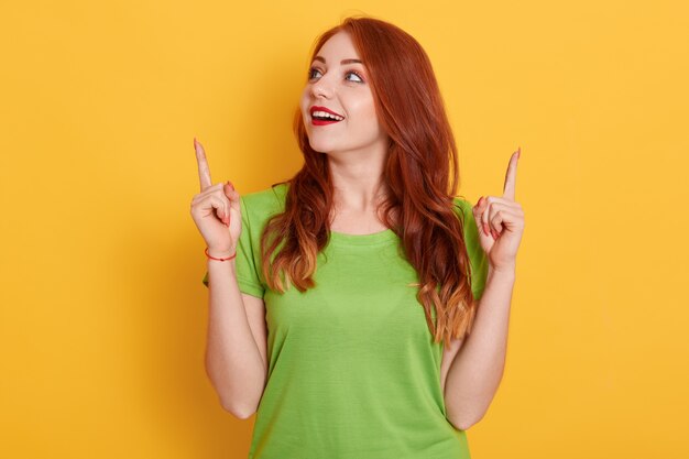 Chica pelirroja feliz sorprendida en camiseta verde casual mirando asombrado y apuntando hacia arriba con los dedos índices, posando aislado