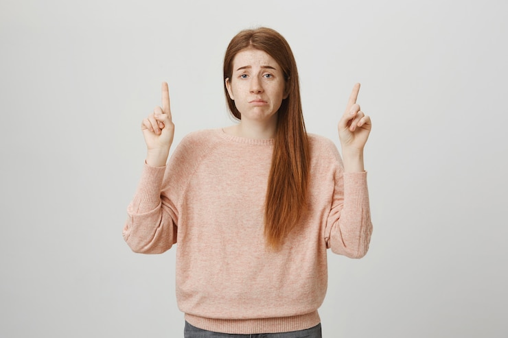 Foto gratuito chica pelirroja molesta sombría haciendo pucheros indecisos, señalando con el dedo hacia arriba