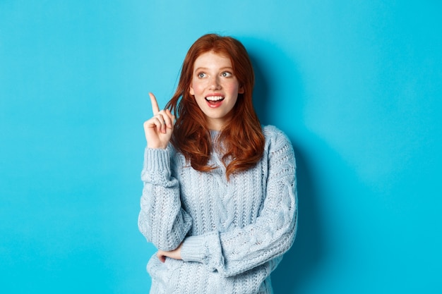 Chica pelirroja pensativa que tiene una idea, levantando el dedo y sonriendo complacida con un buen plan, de pie sobre fondo azul.