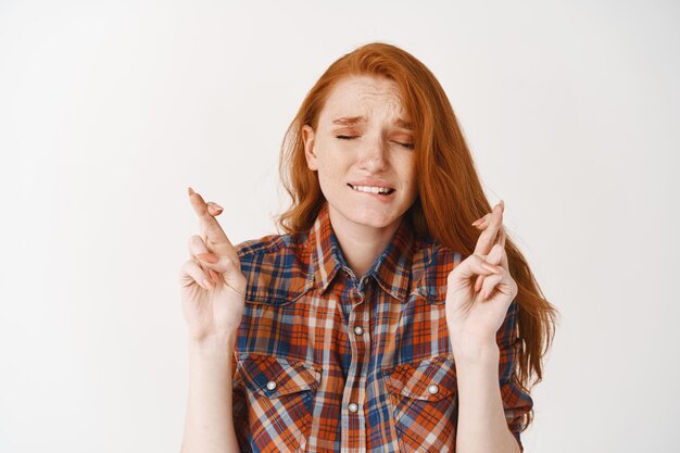 Chica pelirroja preocupada pidiendo un deseo con los dedos cruzados y los ojos cerrados, mordiéndose el labio nerviosamente, de pie sobre una pared blanca