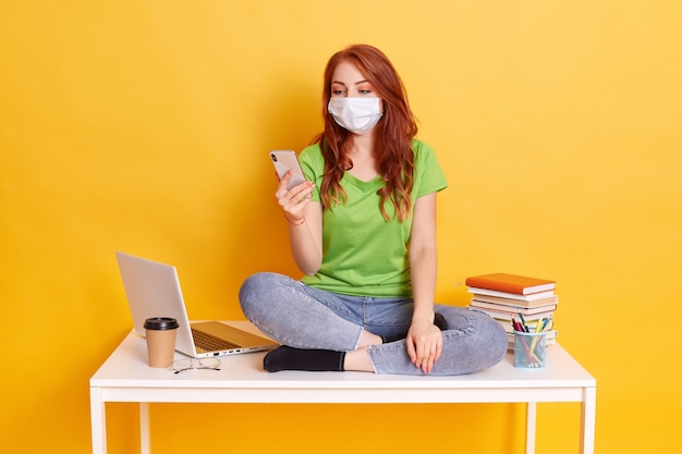 Chica pelirroja de secundaria se sienta en la mesa en posición de loto, usa teléfonos inteligentes, charla con amigos mientras se toma un descanso, usa jeans y camiseta verde, máscara médica aislada sobre fondo amarillo.