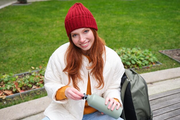 Chica pelirroja sonriente descansa en el parque se sienta en un banco con bebidas de mochila de termo disfruta de una bebida caliente