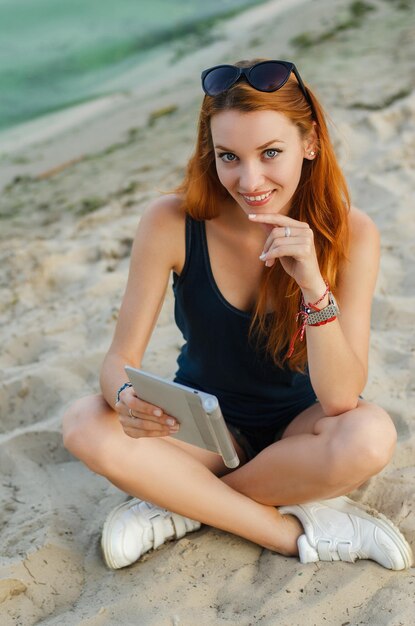 Chica pelirroja sonriente sentada en la playa y sosteniendo una tableta.