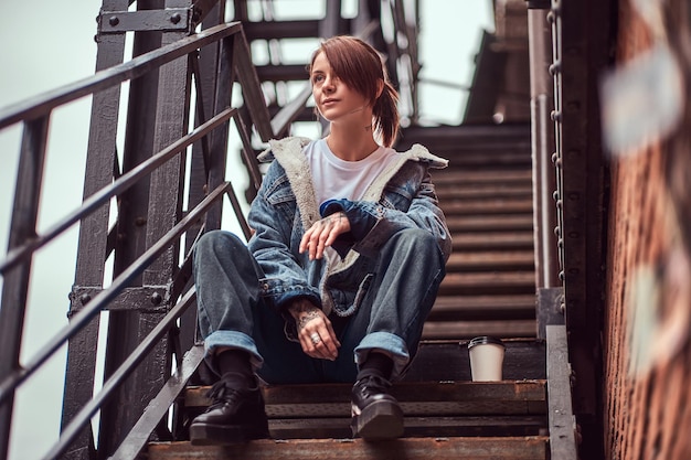 Una chica pelirroja tatuada con ropa de moda sentada en las escaleras afuera.