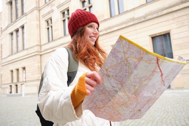 Foto gratuita chica pelirroja turista explora la ciudad mira el mapa de papel para encontrar el camino para los monumentos históricos mujer en