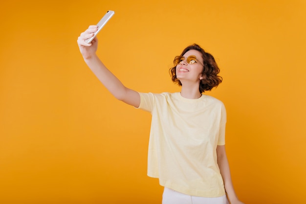 Chica de pelo castaño en camiseta blanca con teléfono para selfie