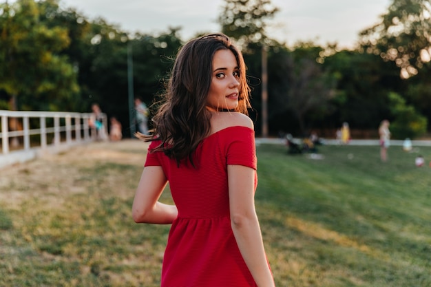 Chica de pelo castaño soñadora mirando por encima del hombro mientras camina en el parque. Retrato de increíble dama caucásica en vestido rojo de pie.