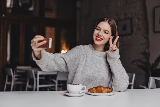 Chica de pelo corto positiva con lápiz labial rojo y sonrisa blanca como la nieve hace selfie en café y muestra signos de paz.
