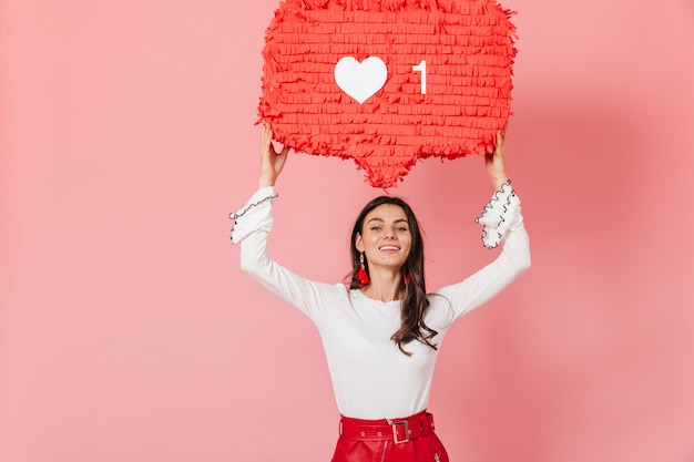 Foto gratuita una chica de pelo largo con aretes rojos y una sonrisa demuestra un enorme me gusta en instagram. retrato de mujer con blusa blanca sobre fondo rosa.