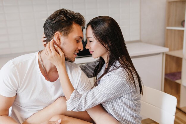 Chica de pelo largo con camisa azul acariciando el rostro de su novio con ternura. Retrato interior de una hermosa dama morena descansando en la cocina con su esposo en buenos días.