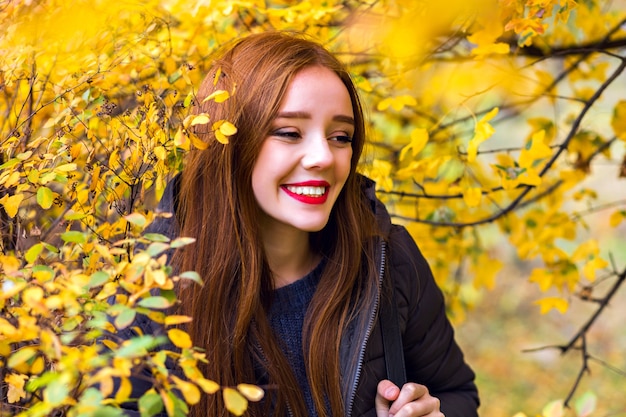 Chica de pelo largo complacida divirtiéndose en el parque con follaje amarillo. Retrato al aire libre de modelo femenino morena riendo mirando a otro lado mientras posa en el bosque