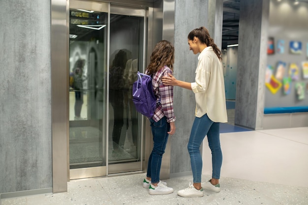 Una chica de pelo largo con una mochila y una sonriente y complacida mujer de pelo oscuro parada ante las puertas cerradas del ascensor