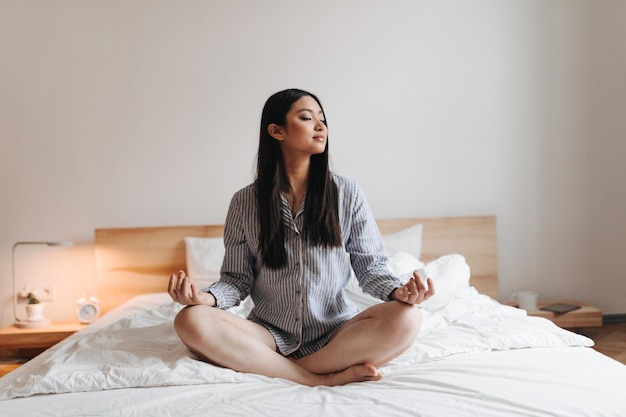 Chica de pelo largo pacificada en pijama está meditando Instantánea de Asia en un dormitorio brillante