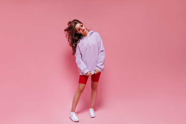 Chica de pelo largo en pantalones cortos de ciclismo y poses con capucha de gran tamaño púrpura sobre fondo rosa Retrato de mujer encantadora en gafas de sol rojas en aislado