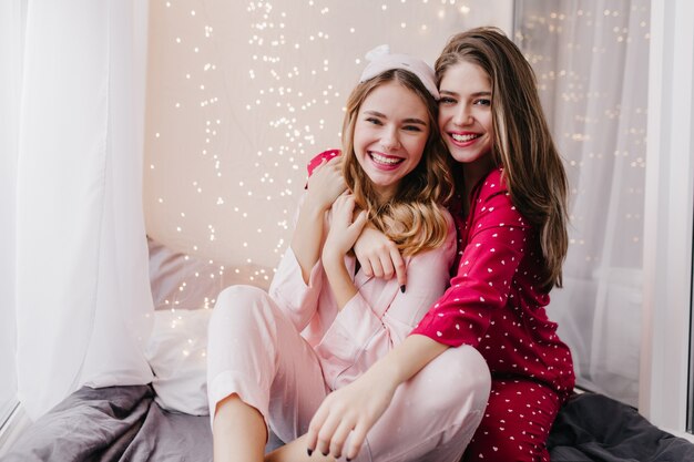 Chica de pelo largo posando emocionalmente junto a la hermana en la cama. Filmación en interiores de espectaculares modelos femeninas en pijama sentado en el dormitorio.