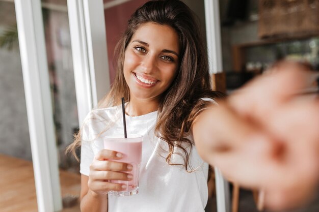 Chica de pelo largo con sonrisa blanca como la nieve hace selfie sosteniendo un vaso de batido