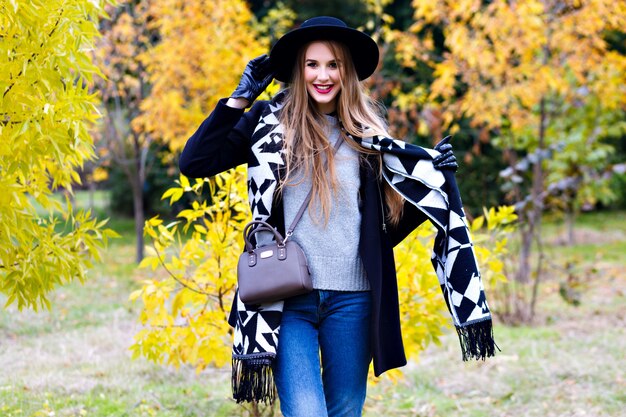 Chica de pelo largo viste jeans jugando con su bufanda en el parque de otoño. Mujer joven atractiva con sombrero negro elegante pasar tiempo en el bosque y riendo.