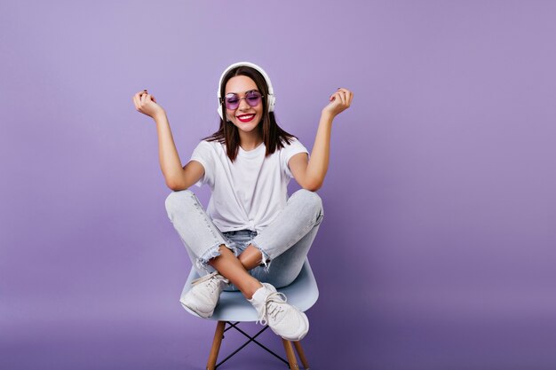 Chica de pelo oscura inspirada sentada en una silla y riendo. Foto interior de sonriente increíble modelo femenino en ropa blanca y auriculares.