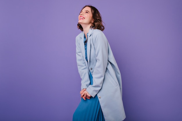 Chica de pelo oscura positiva sonriendo durante la sesión de fotos en abrigo azul. Increíble mujer rizada que expresa emociones positivas en la pared púrpura.