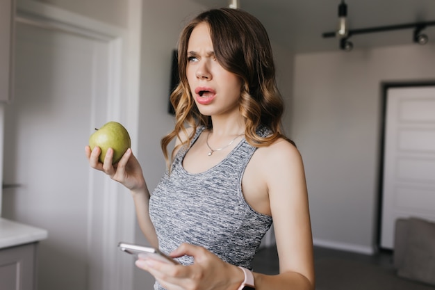 Chica de pelo oscuro con maquillaje nude posando en casa con smartphone. Infeliz mujer joven con manzana y teléfono de pie con la boca abierta.