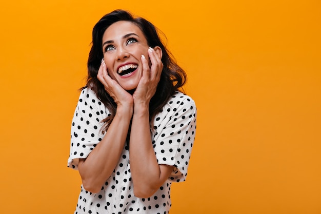 Chica de pelo oscuro mira soñadoramente sobre fondo naranja. Mujer sorprendida con ojos verdes en blusa de lunares blancos sonriendo y posando.