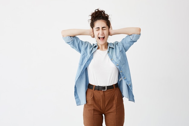 Chica con el pelo oscuro en moño, lleva camisa vaquera y pantalones marrones, se tapa las orejas con las manos, hace una mueca, no quiere escuchar chismes. La mujer enojada no puede soportar el sonido fuerte, grita con los ojos cerrados